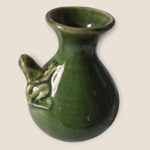 May include: A small, green ceramic vase with a frog-shaped spout and a frog figurine on the side. The vase has a rounded body and a wide opening. The frog is detailed with textured skin and outstretched limbs.