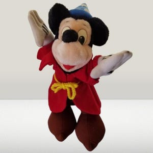 May include: A plush Mickey Mouse toy dressed as a sorcerer. He is wearing a red robe with a yellow sash, a blue pointed hat, and white gloves. He is standing with his arms outstretched.