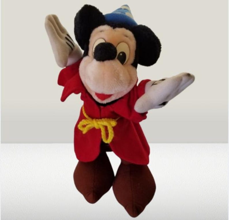 May include: A plush Mickey Mouse toy dressed as a sorcerer. He is wearing a red robe with a yellow sash, a blue pointed hat, and white gloves. He is standing with his arms outstretched.