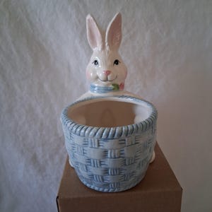 May include: A ceramic Easter bunny planter with a basket design. The bunny is white with pink inner ears, a pink nose, and blue eyes. The basket is light blue with a woven pattern. A blue scarf and a pink flower adorn the bunny's neck.