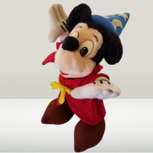 May include: A plush Mickey Mouse dressed as a sorcerer, wearing a red robe, a blue and white hat, and yellow shoes. He is holding his right hand up in a magical gesture.