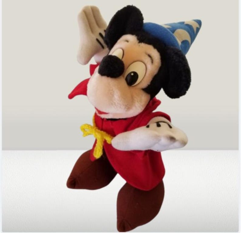 May include: A plush Mickey Mouse dressed as a sorcerer, wearing a red robe, a blue and white hat, and yellow shoes. He is holding his right hand up in a magical gesture.