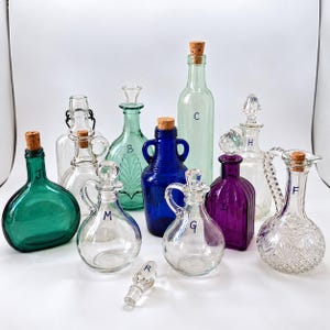 May include: A collection of various glass bottles and decanters in clear, green, blue, and purple hues. Several feature cork stoppers, while others have glass stoppers. Some bottles are marked with letters including J, B, C, H, M, G, I, and F.