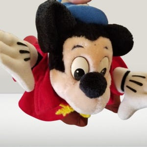 May include: A plush Mickey Mouse toy wearing a red and gold robe and a blue hat. The toy has a white face with black eyes and a black nose. The toy is in a flying pose with its arms outstretched.