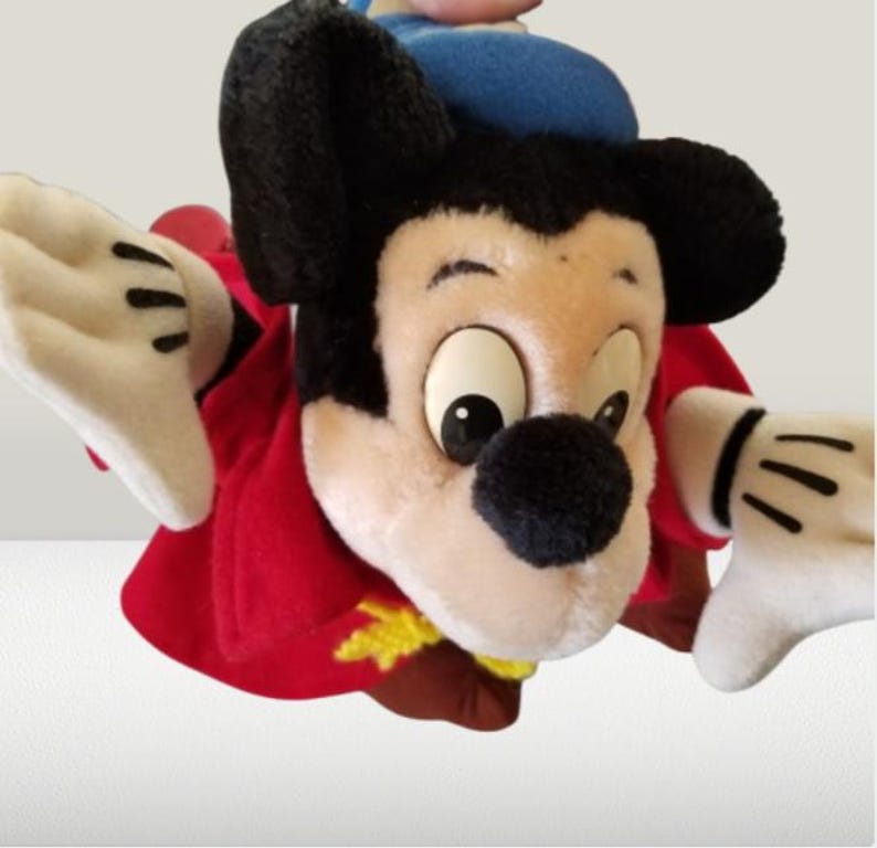 May include: A plush Mickey Mouse toy wearing a red and gold robe and a blue hat. The toy has a white face with black eyes and a black nose. The toy is in a flying pose with its arms outstretched.