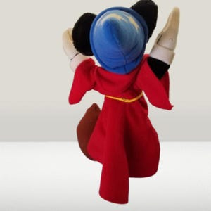 May include: A plush toy of Mickey Mouse dressed as a sorcerer. He is wearing a red robe with a yellow belt, a blue hat with stars, and white gloves.
