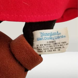 May include: A brown and black plush Mickey Mouse toy wearing a red hat with a white tag that reads "Disneyland Walt Disney World&reg; DISNEY ALL NEW MATERIALS POLYESTER FIBERS WE 1525. PA 1378:OHIO PA-1378 MASS T-618 CARASA TRADING CORP BURBANK, CA. PRODUCT OF KOREA MER-100"