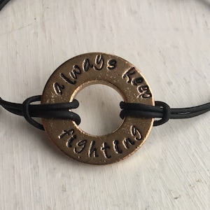 Może przedstawiać: Bransoletka z zawieszką w kształcie podkładki w kolorze brązu z czarnym napisem "always keep fighting". Zawieszka jest połączona z czarną bransoletką ze sznurka.