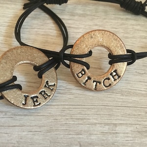 Könnte beinhalten: Zwei kupferfarbene Unterlegscheiben-Armbänder mit schwarzen Kordeln. Eine Scheibe trägt die Aufschrift "JERK", die andere "BITCH". Die Armbänder werden auf einer hellen Holzmaserungsoberfläche präsentiert.