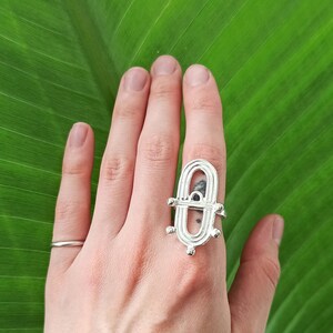May include: Silver-toned ring with an elongated oval design, featuring a central open space and decorative elements. The ring is worn on a hand, with a simple band ring on another finger. The background is a large green leaf.