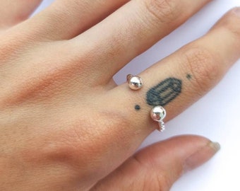 Puede incluir: Un anillo plateado con dos adornos esféricos. El anillo se lleva en un dedo, con una esfera en el lateral y otra en la parte superior. El diseño del anillo es minimalista y moderno.