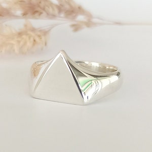 Op de afbeelding: Een zilveren ring met een geometrisch piramide-ontwerp. De ring ligt op een witte ondergrond met een vage achtergrond van gedroogde planten. De piramidevorm van de ring is het middelpunt en reflecteert licht.