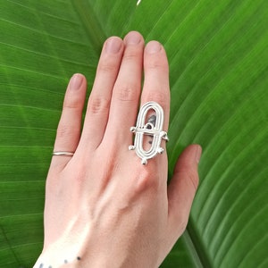 May include: A silver ring with an open, geometric design is displayed on a hand against a large, green leaf. The ring has an elongated oval shape with a central bar and decorative elements. A simple silver band ring is also visible.