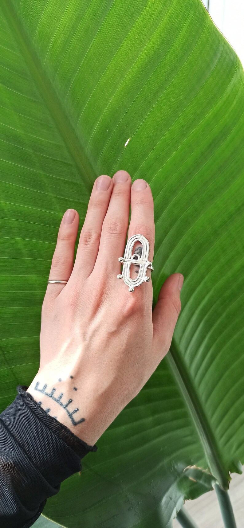 May include: A silver ring with an open, geometric design is displayed on a hand against a large, green leaf. The ring has an elongated oval shape with a central bar and decorative elements. A simple silver band ring is also visible.