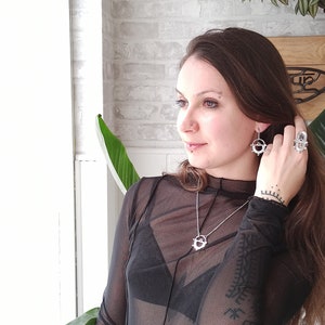 May include: A woman wearing silver jewelry, including a necklace, earrings, and a ring. The jewelry features a circular design. She is wearing a sheer black top and dark blue jeans. The background includes a white brick wall and plants.