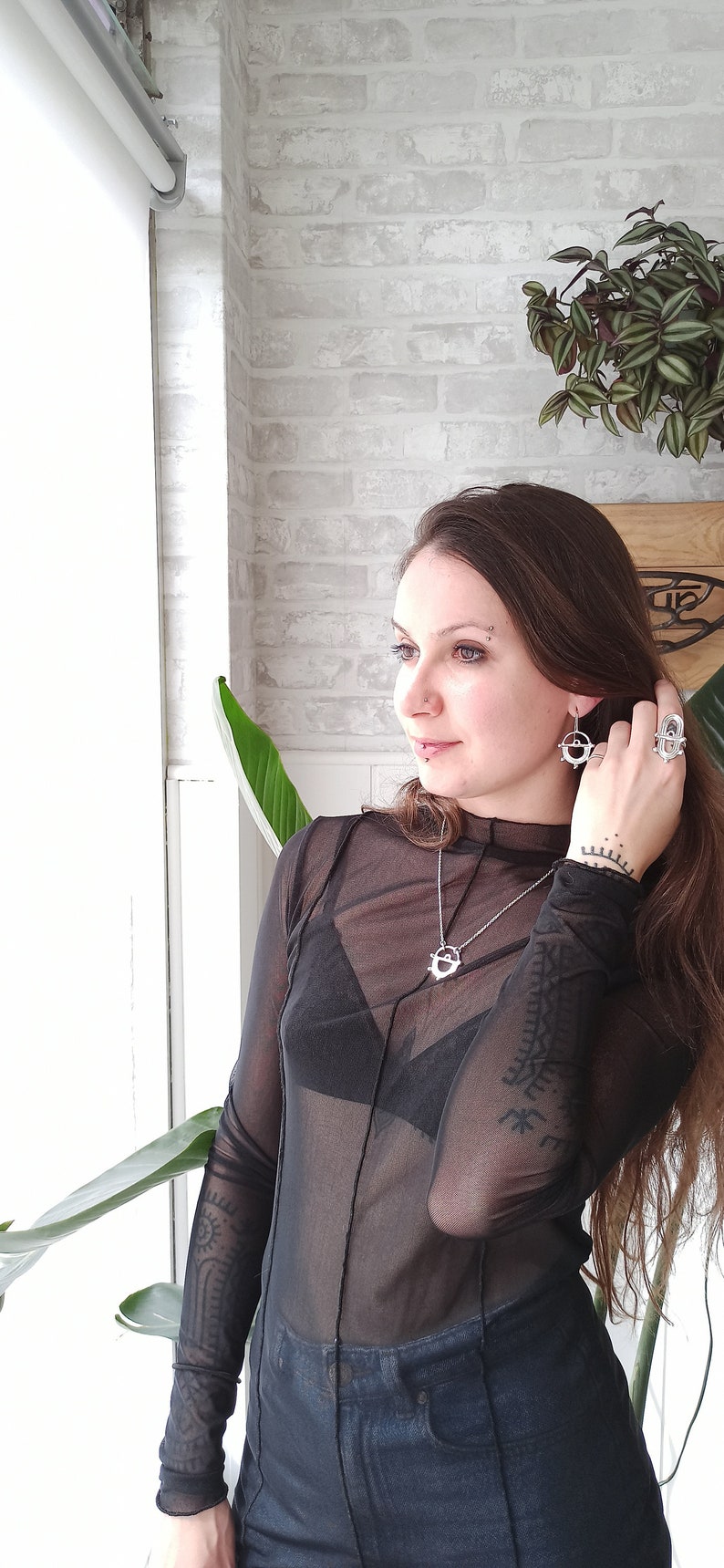 May include: A woman wearing silver jewelry, including a necklace, earrings, and a ring. The jewelry features a circular design. She is wearing a sheer black top and dark blue jeans. The background includes a white brick wall and plants.