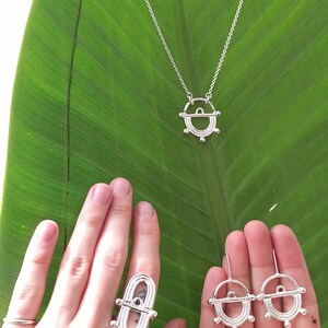 May include: Silver-toned jewelry set including a necklace, ring, and earrings, displayed on a large green leaf. The necklace has a pendant with a circular design. The ring and earrings share a similar design. The jewelry is likely made of sterling silver.