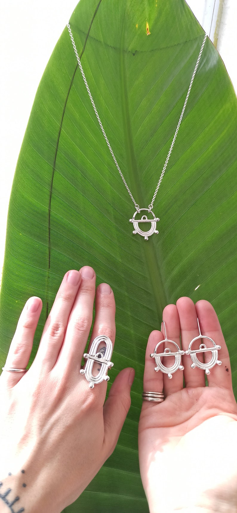 May include: Silver-toned jewelry set including a necklace, ring, and earrings, displayed on a large green leaf. The necklace has a pendant with a circular design. The ring and earrings share a similar design. The jewelry is likely made of sterling silver.