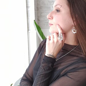 May include: A silver ring and matching earrings with geometric designs. The ring is on a finger, and the earrings are dangling. The person is wearing a sheer black top and a silver necklace. The background includes a white wall and a plant.