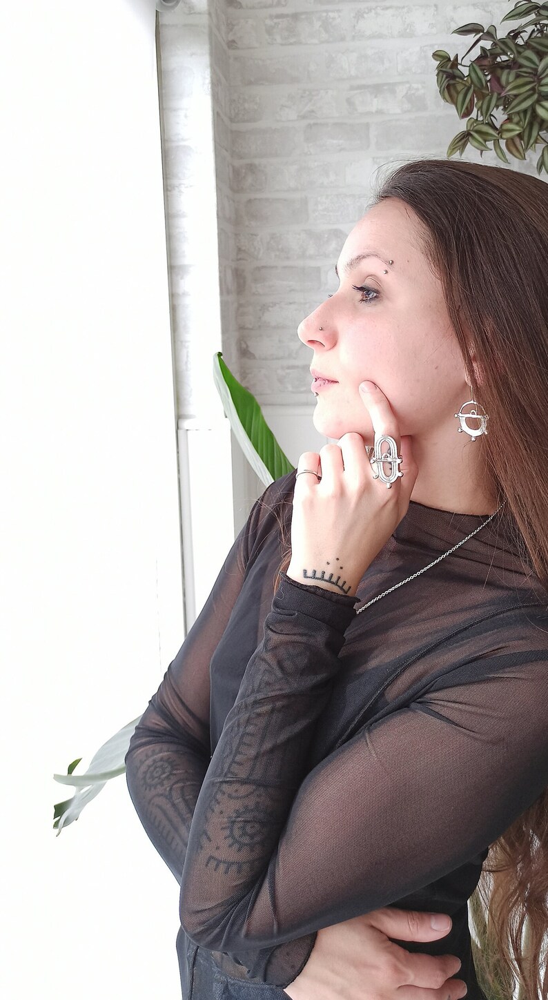 May include: A silver ring and matching earrings with geometric designs. The ring is on a finger, and the earrings are dangling. The person is wearing a sheer black top and a silver necklace. The background includes a white wall and a plant.