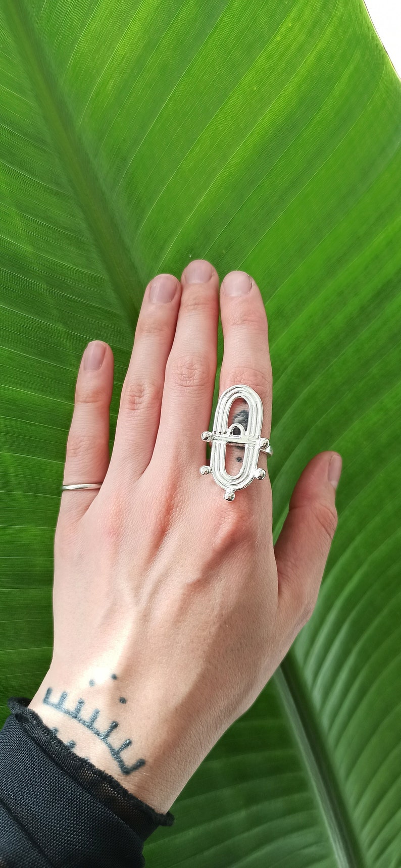 May include: Silver-toned ring with an elongated oval design, featuring a central open space and decorative elements. The ring is worn on a hand, with a simple band ring on another finger. The background is a large green leaf.