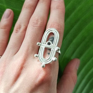 May include: A silver ring with an open, geometric design featuring an oval shape with a small black stone. The ring is worn on a hand against a green leaf background.