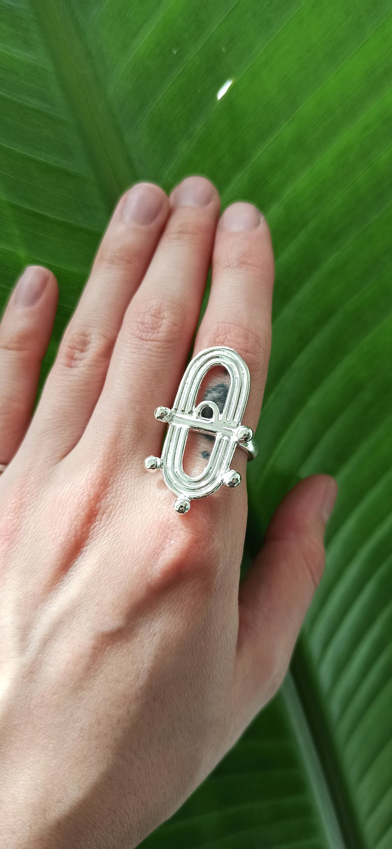 May include: A silver ring with an open, geometric design featuring an oval shape with a small black stone. The ring is worn on a hand against a green leaf background.