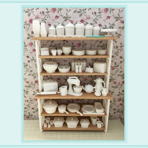 May include: A white kitchen shelving unit with four shelves filled with white dishes, bowls, canisters, and kitchen tools. The shelving unit is against a floral wallpaper background.