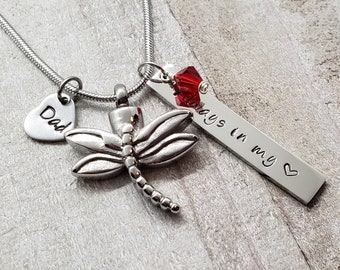 May include: Silver necklace with a dragonfly pendant, a heart charm with the word "Dad" engraved on it, and a bar charm with the words "Always in my heart" engraved on it. A red crystal is attached to the bar charm.