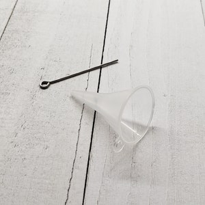 K&ouml;nnte beinhalten: Ein wei&szlig;er Plastik-Trichter mit einem kleinen Griff an der Seite und einem silbernen Metalldraht mit einer Schlaufe an einem Ende, auf einer wei&szlig;en Holzoberfl&auml;che.