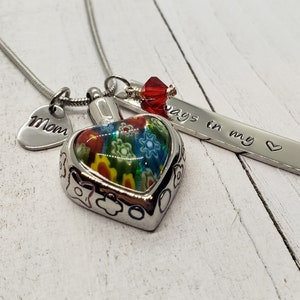 May include: A silver necklace with a heart-shaped pendant featuring a colorful glass design. The necklace also includes a small heart charm with the word "Mom" engraved on it and a rectangular charm with the words "Always in my heart" engraved on it.