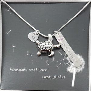 K&ouml;nnte beinhalten: Eine silberne Halskette mit einem Herz-Anh&auml;nger, auf dem "Mom" steht, einem Schildkr&ouml;ten-Anh&auml;nger und einem Barren-Anh&auml;nger, auf dem "Always in my heart" steht. Die Halskette befindet sich in einer schwarzen Schachtel mit einem wei&szlig;en L&ouml;wenzahn und den Worten "handmade with love" und "Best Wishes".