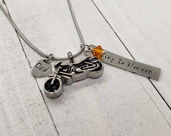 May include: Silver necklace with a motorcycle charm, a heart charm with the word "Dad" engraved on it, and a bar charm with the words "Riding In Heaven". The necklace also has a small orange crystal charm.