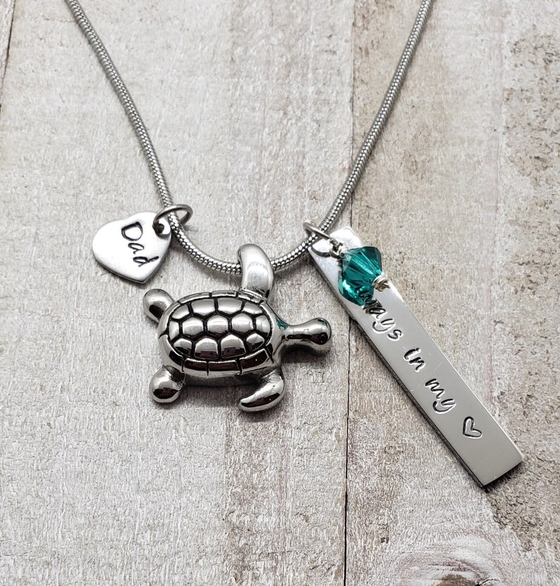 K&ouml;nnte beinhalten: Eine silberne Halskette mit einem Herz-Anh&auml;nger, auf dem "Dad" steht, einem kleinen Schildkr&ouml;ten-Anh&auml;nger und einem rechteckigen Anh&auml;nger mit dem Text "Always in my heart" und einem t&uuml;rkisfarbenen Kristall-Anh&auml;nger.