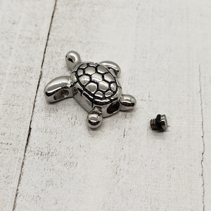 K&ouml;nnte beinhalten: Ein silberner Schildkr&ouml;ten-Krempendant mit einem Schraubverschluss. Der Anh&auml;nger ist offen und der Deckel ist separat.
