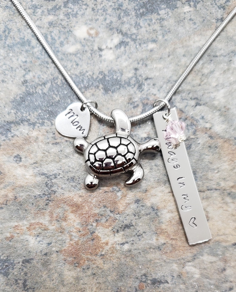K&ouml;nnte beinhalten: Silberne Halskette mit einem Schildkr&ouml;ten-Anh&auml;nger, einem Herz-Anh&auml;nger mit der Gravur "Mom" und einem Barren-Anh&auml;nger mit der Gravur "Always in my heart".