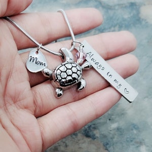 K&ouml;nnte beinhalten: Silberne Halskette mit einem Schildkr&ouml;tenanh&auml;nger, einem Herzanh&auml;nger mit der Aufschrift "Mom" und einem Barrenanh&auml;nger mit der Aufschrift "Always in my heart".