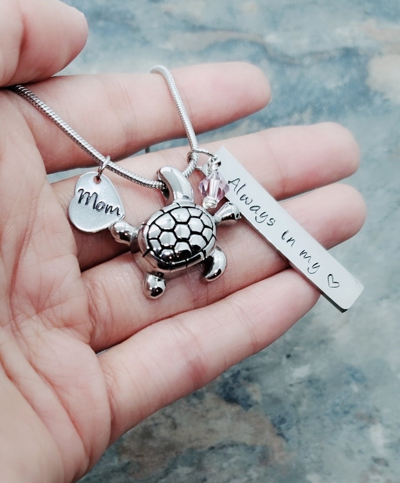 K&ouml;nnte beinhalten: Silberne Halskette mit einem Schildkr&ouml;tenanh&auml;nger, einem Herzanh&auml;nger mit der Aufschrift "Mom" und einem Barrenanh&auml;nger mit der Aufschrift "Always in my heart".