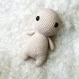 May include: A crocheted doll in a light beige color with black button eyes. The doll has a rounded head, small arms, and stubby legs. The doll is lying on a white, fluffy surface.