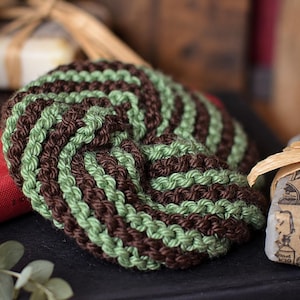 May include: A round, crocheted scrubby featuring a spiraling pattern of alternating dark brown and light sage green yarn. The textured, circular item is constructed with thick, durable stitches that create a dense, raised surface throughout.