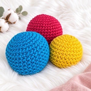 May include: Three spherical objects made of crocheted yarn in solid colours of bright blue, deep red, and vibrant yellow. The textured, hand-stitched yarn balls are arranged in a cluster, showcasing the intricate loop patterns of the crochet work.