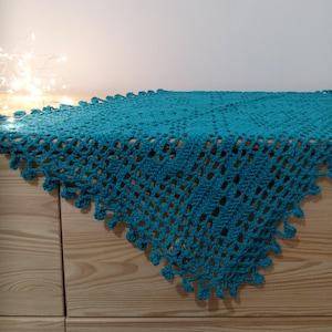 Può includere: Un runner da tavolo all'uncinetto color turchese con un delicato motivo traforato. Il runner è appoggiato su una superficie in legno.