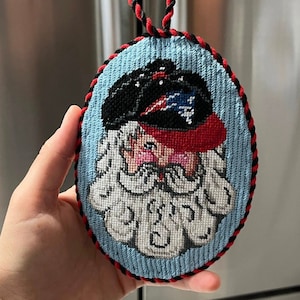 May include: A hand-stitched Christmas ornament featuring a Santa Claus wearing a black and red hat with a white star. The ornament is oval-shaped and has a red and black trim.