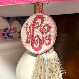 May include: A white lampshade with a pink fabric cover. A pink and white embroidered round ornament with the word "Hope" stitched in pink hangs from the lampshade with a cream tassel.