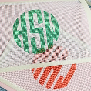 May include: Two white canvas embroidery hoops with green and orange embroidery designs. The top hoop has a green monogram design and the bottom hoop has an orange striped design.