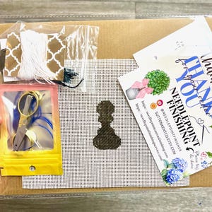 May include: A needlepoint kit with a brown background, a white grid, a brown silhouette of a woman, and a thank you card with blue flowers. The kit includes white thread, a small pair of scissors, and a small bag of beads.