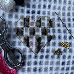 May include: A checkerboard heart needlepoint design in black and white, outlined in gold thread, on white canvas. Accessories include scissors, thread, a needle, and gold charms with the year 2021.