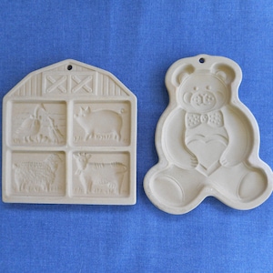 Pode incluir: Dois cortadores de biscoitos, um com a forma de um celeiro com quatro recortes de animais e o outro com a forma de um urso de peluche segurando um coração. Ambos são feitos de um material cerâmico de cor marrom claro.