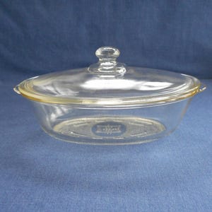 May include: A clear glass oval baking dish with a lid. The dish has a raised rim and a textured bottom. The lid has a knob on top.