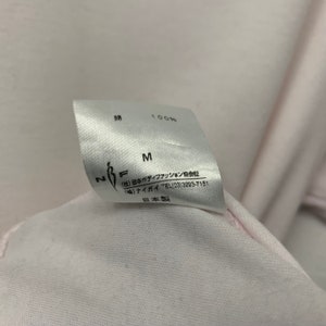 May include: Close-up of a white clothing tag with Japanese text and the letters "M" and "N F". The tag indicates 100% cotton. The text includes the name of a Japanese body fashion association and a phone number.